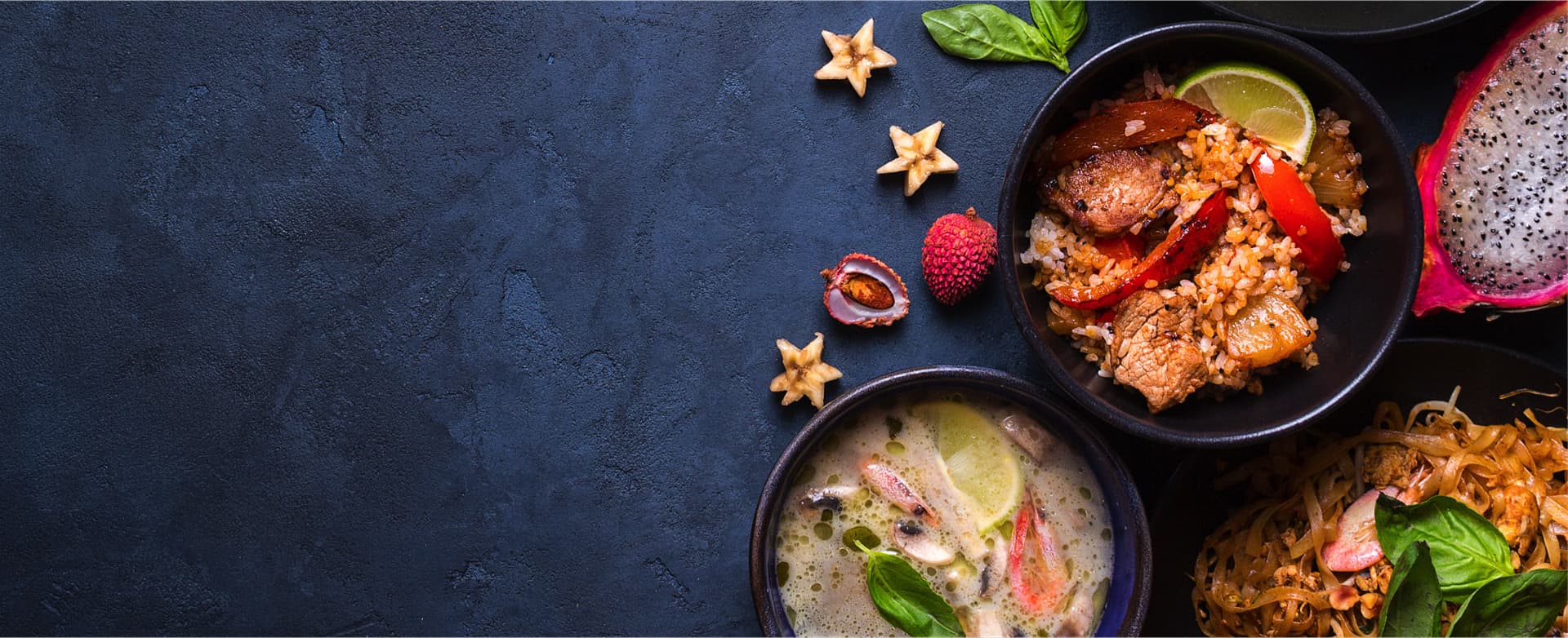 A photo showing an assortment of dishes from Asian cuisine including a fish and vegetables soup, fried rice with grilled pork and peppers, sauted noodles with cabbage and psices, and a pice of dragon fruit cut in half, all arranged on a concrete kitchen counter.